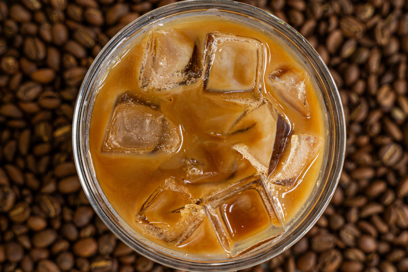 Glass of iced coffee with ice cubes on a bed of roasted coffee beans, showcasing Heyday Eatery’s refreshing cold brew made from premium roasted beans.