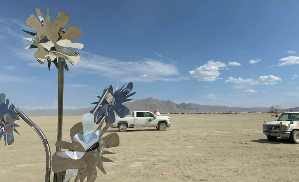 Metal sunflower sculpture by Heyday Eatery shining in the desert at Burning Man festival, with pickup trucks and distant mountains under a clear blue sky.