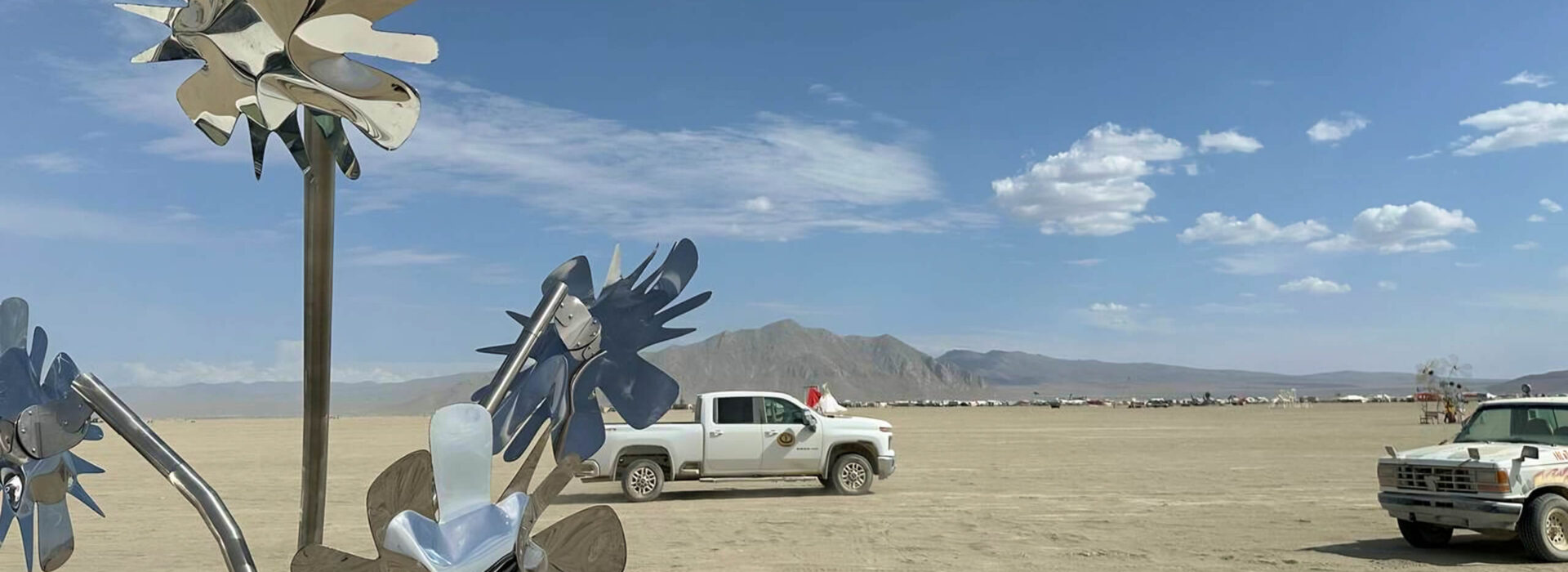 Metal sunflower sculpture by Heyday Eatery shining in the desert at Burning Man festival, with pickup trucks and distant mountains under a clear blue sky.