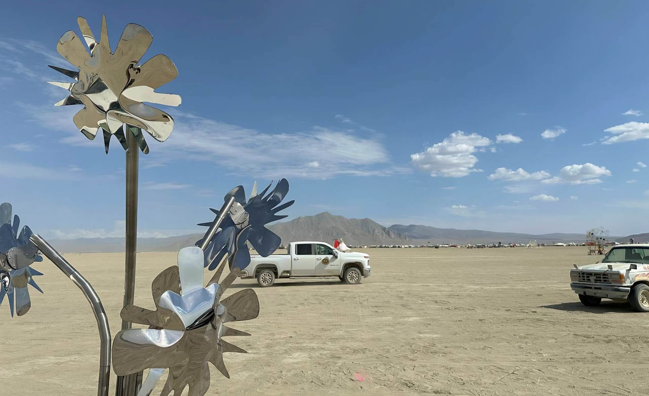 Metal sunflower sculpture by Heyday Eatery shining in the desert at Burning Man festival, with pickup trucks and distant mountains under a clear blue sky.